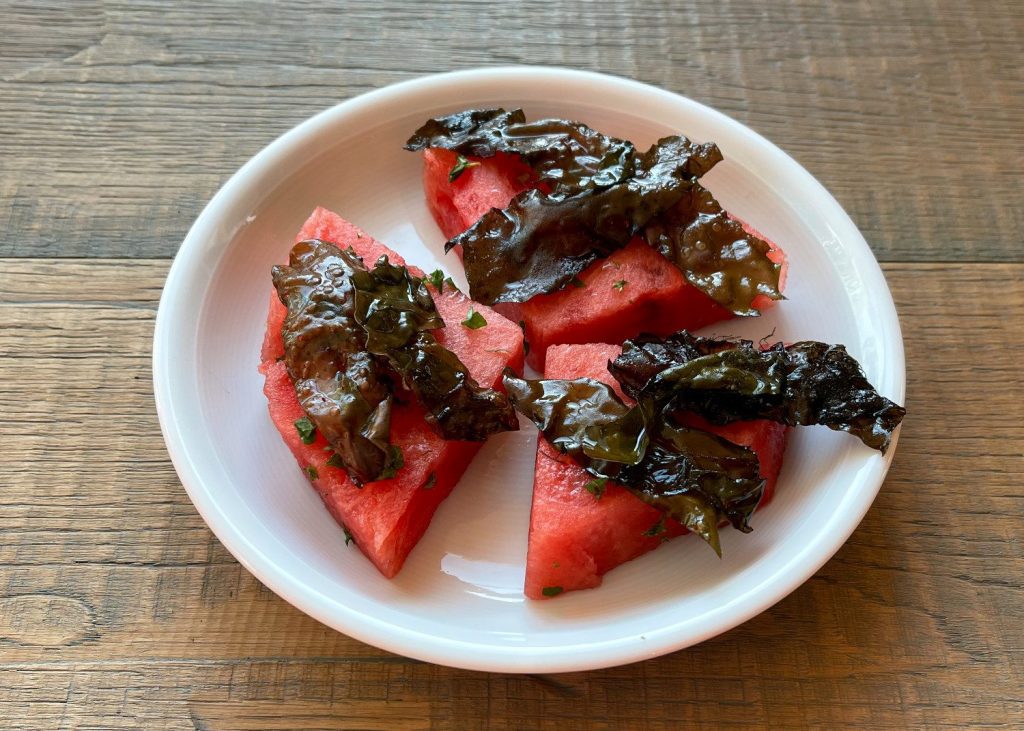 Drei Stücke von Wassermelone belegt mit Dulse auf einem weißen Teller, so geht Kochen mit Algen sehr einfach
