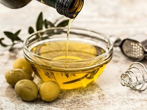 Close-up of olive oil being poured into a glass bowl surrounded by fresh olives and kitchen tools.